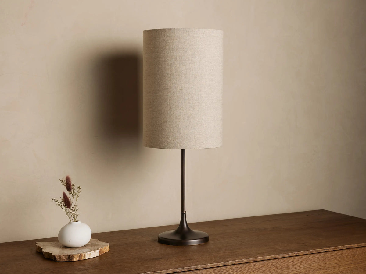 Lorelei Table Lamp with a dark bronze base and stem, and an illuminated cylindrical flax linen shade on a wood table.