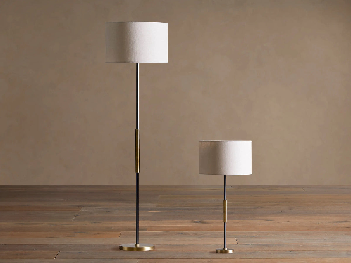 Close-up of Ardea Floor Lamp's base, showing a matte black pole entering a brushed antique brass cylinder on a blurred wood floor.