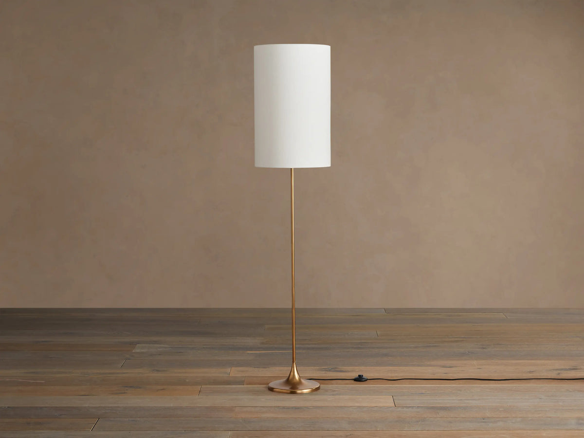 Lorelei Floor Lamp with antiqued brass finish, tall cylindrical white linen shade, standing on a wooden floor.