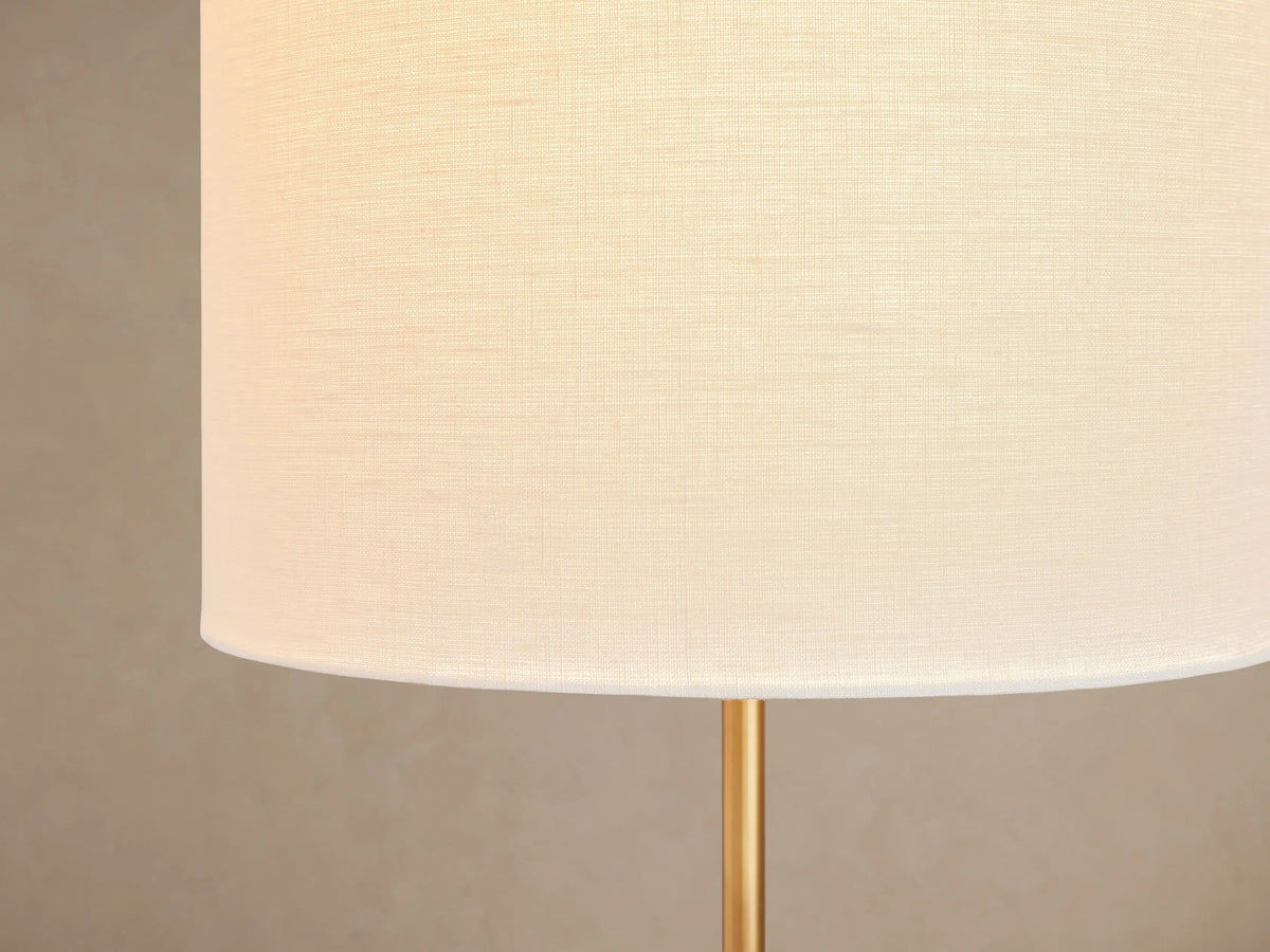 Antiqued brass Lorelei Floor Lamp, slender stem, round base, and tall white linen cylindrical shade on wood floor.