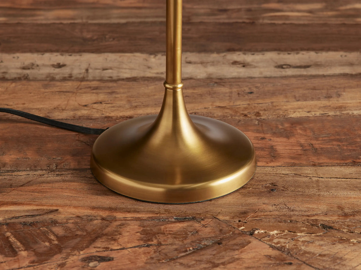 Lorelei Table Lamp, antique brass with a slender stand, round base, and a tall, cylindrical white linen shade, on wood.