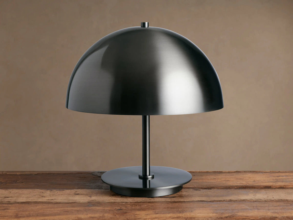 Malcom Table Lamp: dark brushed pewter dome shade, thin stem, and round base on a rustic wooden table.
