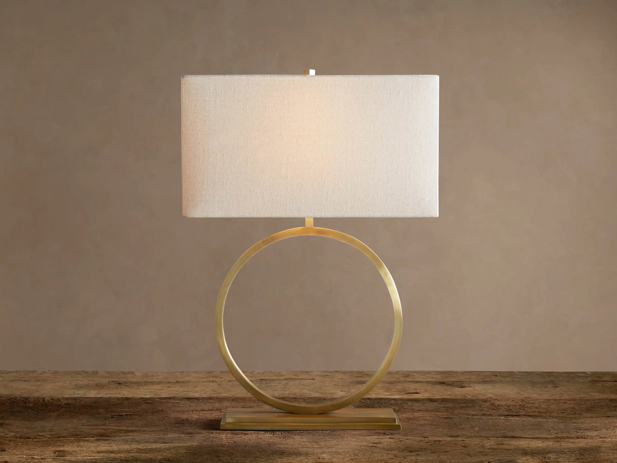 Rhys Table Lamp: Illuminated rectangular light fabric shade atop a gold circular hoop base on a rustic wood surface.