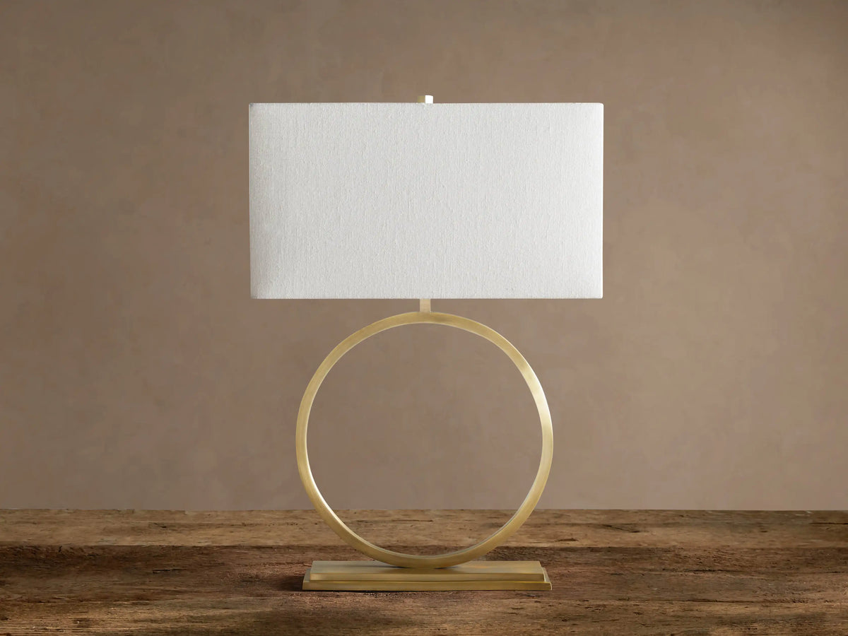 Rhys Table Lamp: Illuminated rectangular light fabric shade atop a gold circular hoop base on a rustic wood surface.