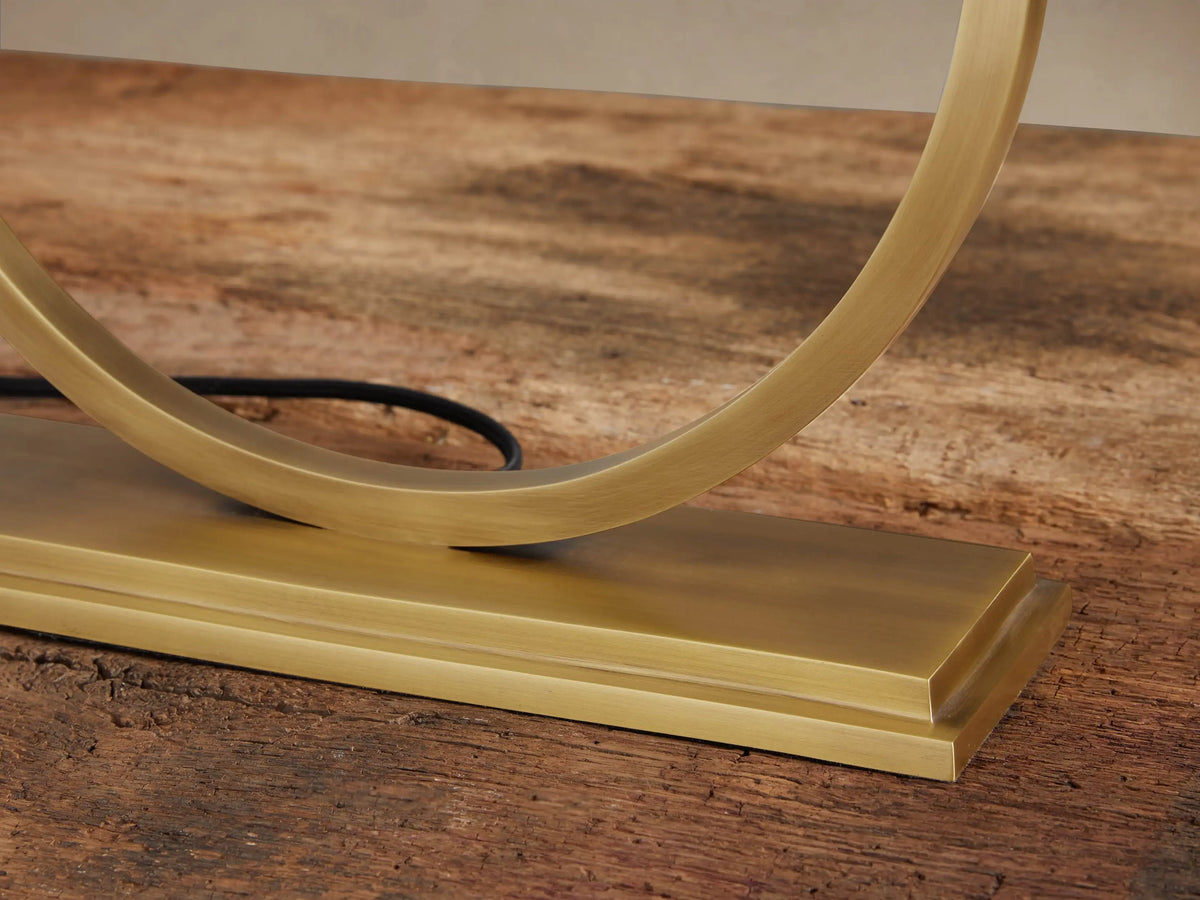 Rhys Table Lamp: White rectangular fabric shade, gold circular metal base, sitting on a rustic wooden table.