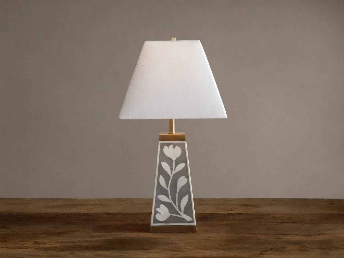 Divya Table Lamp with white fabric shade, gray base with white bone inlay floral motifs, brass trim, on wooden table.