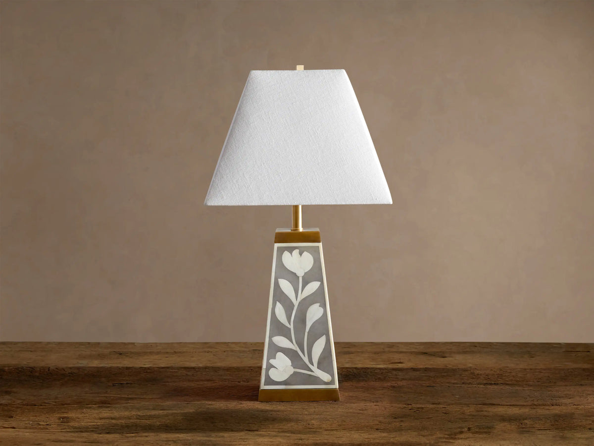 Divya Table Lamp with white fabric shade, gray base with white bone inlay floral motifs, brass trim, on wooden table.