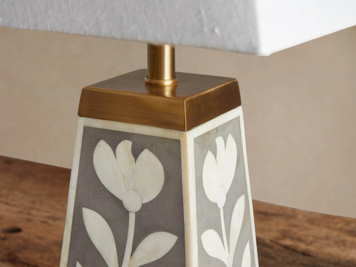 Divya Table Lamp with white trapezoidal shade, brass neck, grey tapered base featuring white bone floral inlay, on wood.