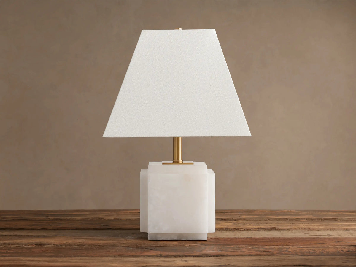 Fatima Table Lamp with a white natural stone base, gold stem, and tapered white linen shade, glowing softly on a wooden table.