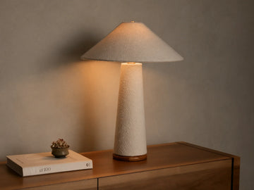 Front view of a lit table lamp featuring a light beige textured cone shade and matching tapered cylindrical base, on a wooden surface.