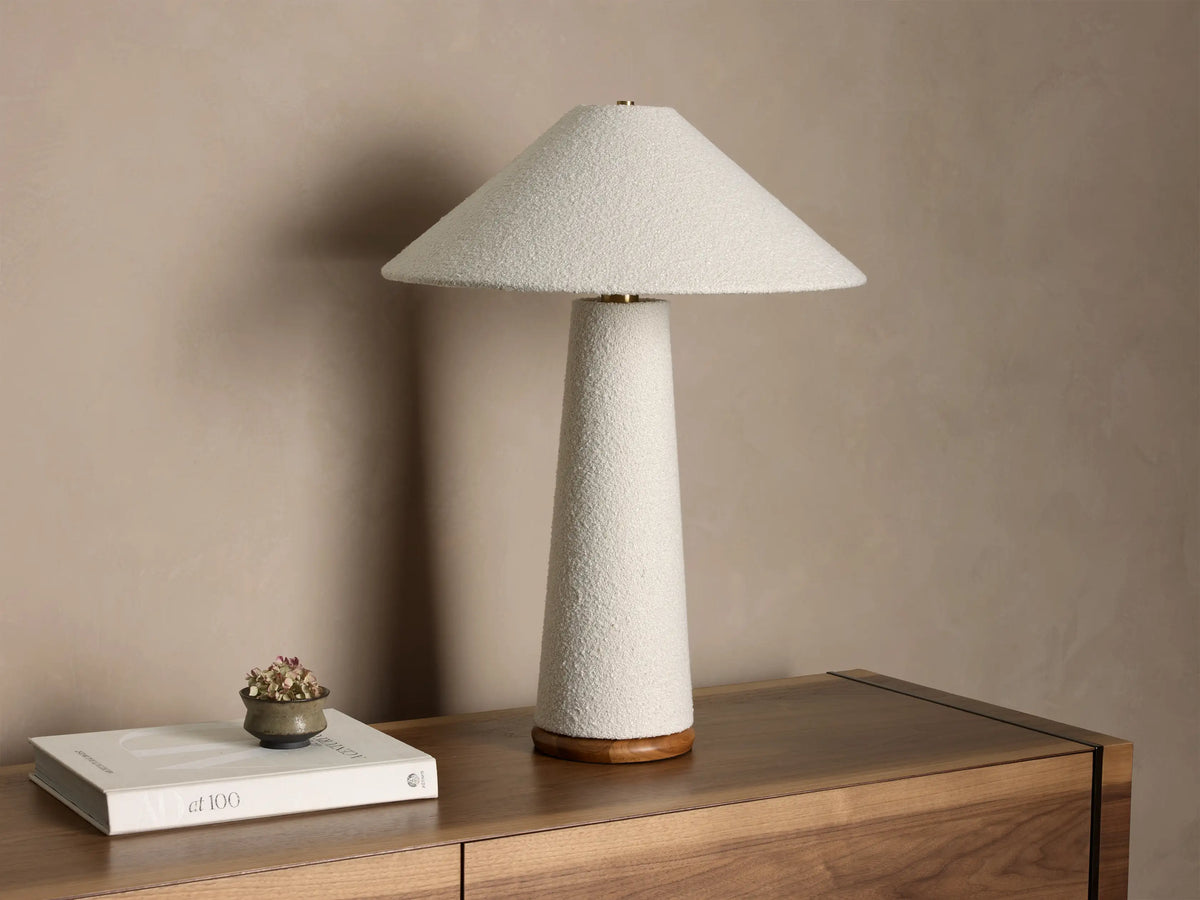 Front view of a lit table lamp featuring a light beige textured cone shade and matching tapered cylindrical base, on a wooden surface.