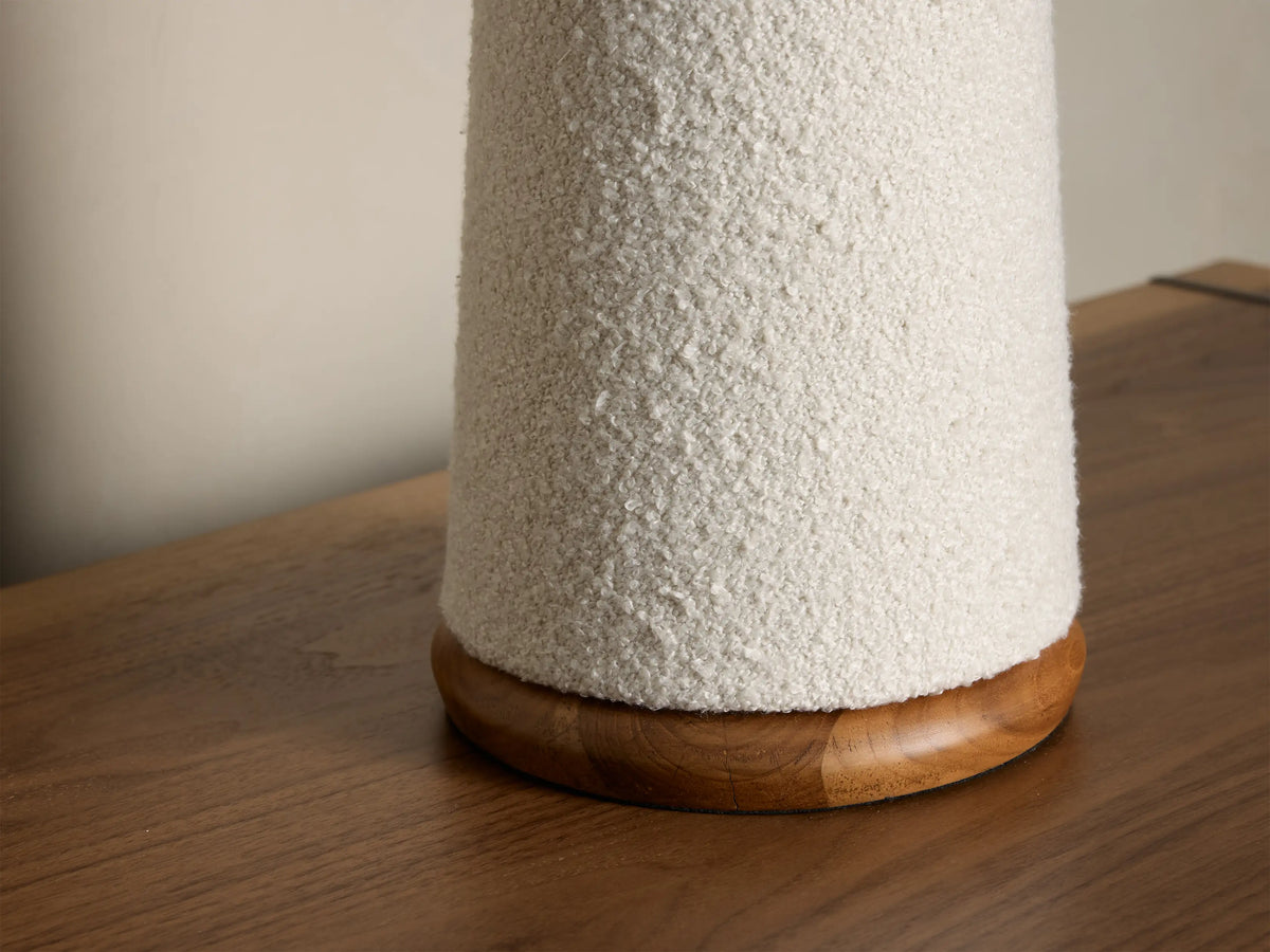 Cream textured table lamp with conical shade, base, brass connector, and wooden foot, on a wooden surface.