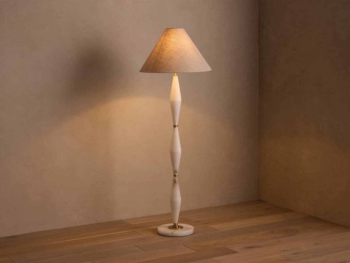 Full view of a floor lamp with a beige conical shade, off-white sculpted stem, and gold accents, casting a warm glow.