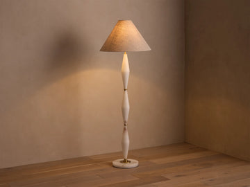 Full view of a floor lamp with a beige conical shade, off-white sculpted stem, and gold accents, casting a warm glow.