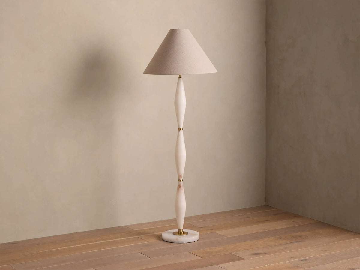 Full view of a floor lamp with a beige conical shade, off-white sculpted stem, and gold accents, casting a warm glow.