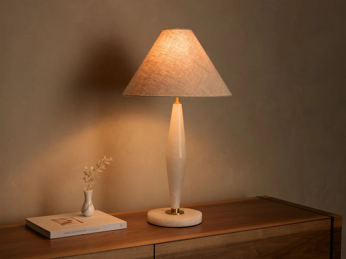 Front view of a lit table lamp with a conical beige fabric shade and a tapered creamy alabaster base with brass details.