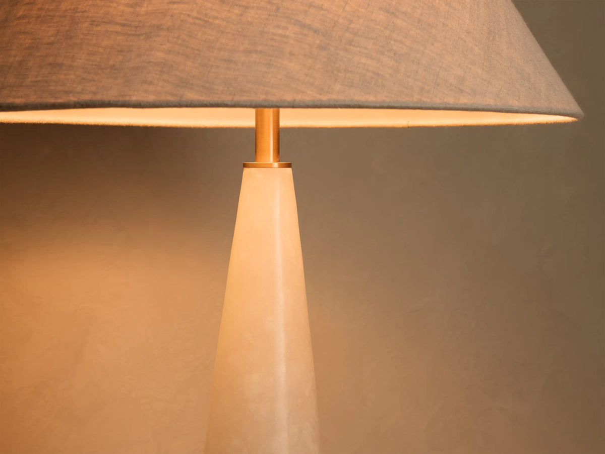 Close-up of a table lamp with a gray fabric lampshade and a gold-toned stem over a light-colored conical base, emitting warm light.