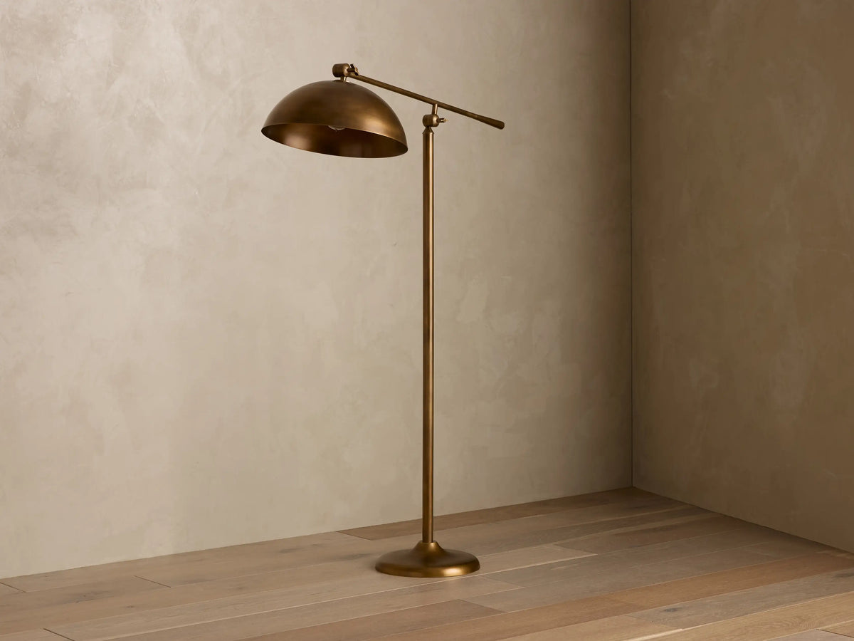 Interior view of a bronze floor lamp with a domed shade angled down, emitting a warm glow, standing on a wooden floor in a room corner.