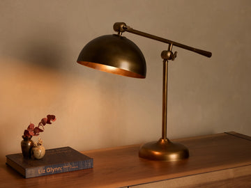 Front view of an illuminated brass industrial-style adjustable table lamp with a domed shade on a wooden surface.
