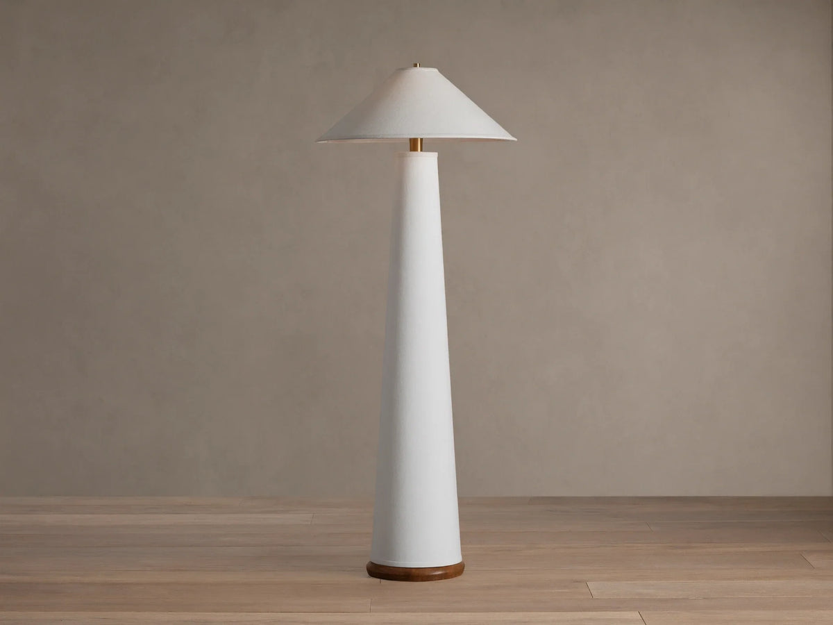 Ombra Floor Lamp: a tall white conical lamp with a white linen shade, teak base, and antiqued brass accents, on a wooden floor.