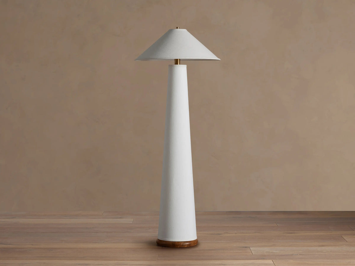 Ombra Floor Lamp: a tall white conical lamp with a white linen shade, teak base, and antiqued brass accents, on a wooden floor.