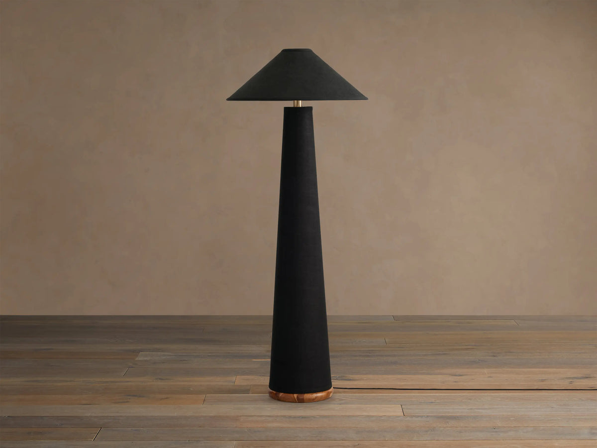 Ombra Floor Lamp. Tall, black linen-wrapped conical lamp with matching shade, brass top accent, and visible teak wood base. Emits warm light.