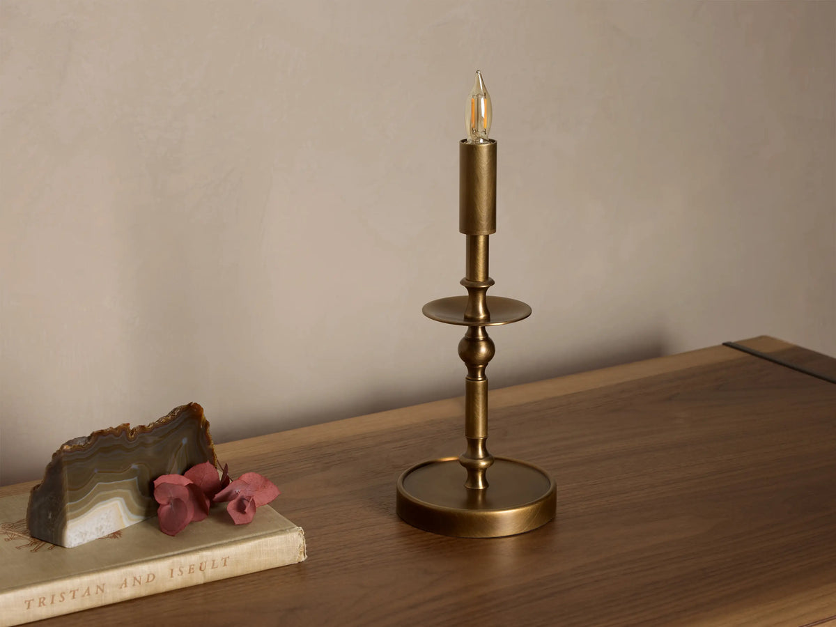 Front view of an antique brass Winslow Candlestick Table Lamp with a slender post, a round base, and a candle-style bulb.