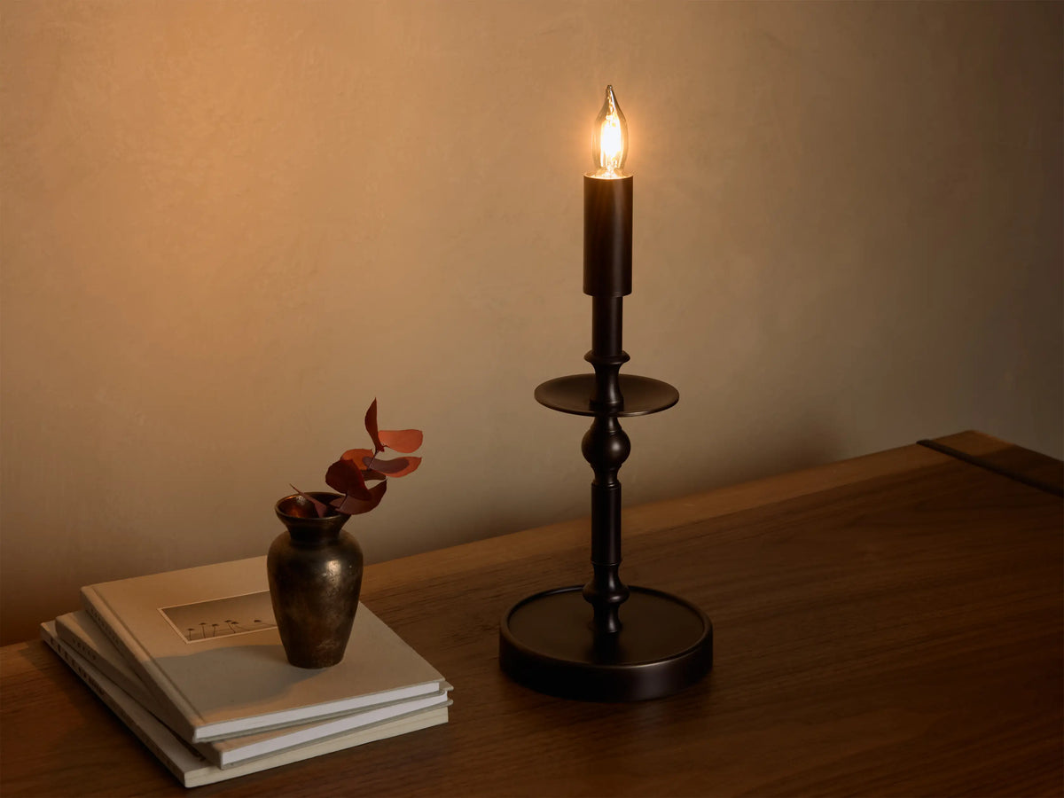 Front view of the illuminated bronze Winslow Candlestick Table Lamp, on a wooden table with books and a small vase.
