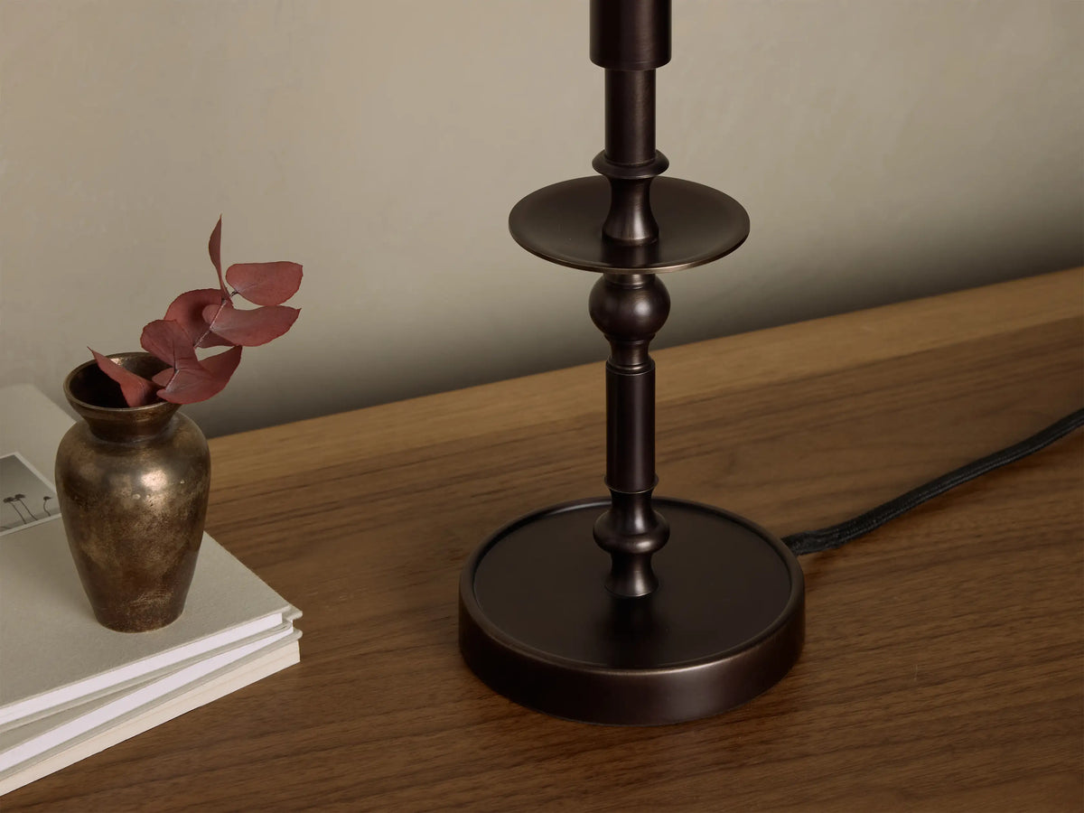 Front view of the illuminated bronze Winslow Candlestick Table Lamp, on a wooden table with books and a small vase.