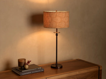 Illuminated Winslow Table Lamp with a bronze-finish iron base and a light brown patterned fabric drum shade on a wooden surface.