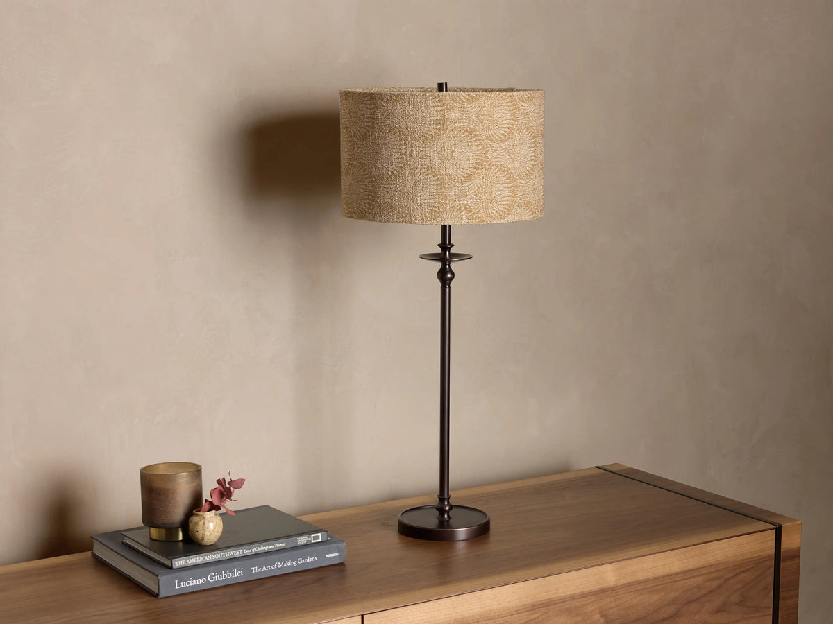 Front view of the Winslow Table Lamp with an antique bronze finish base and a patterned beige fabric shade on a wooden credenza.