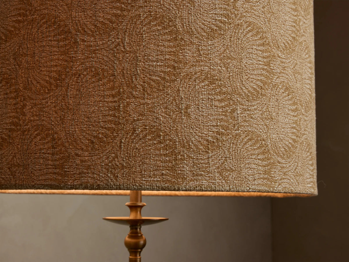 Close-up of the Winslow Floor Lamp's tan fabric shade with subtle lighter leaf pattern, illuminated from within, over an antique brass base.