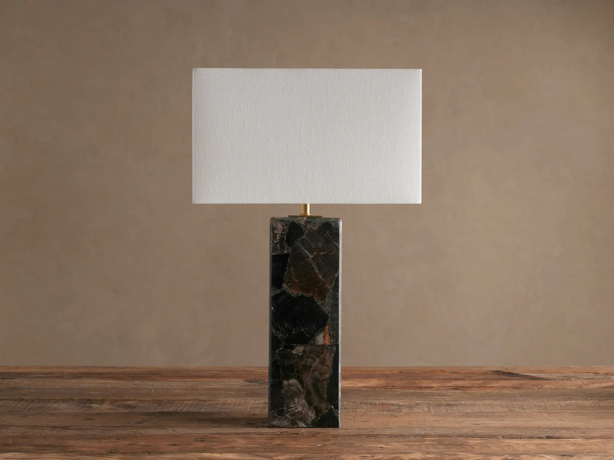 Oren Table Lamp with rectangular white linen shade on a tall dark stone base, standing on a rustic wooden table.