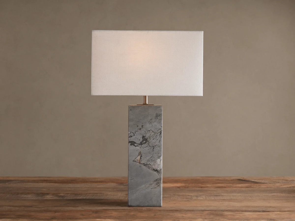 Oren Table Lamp: Rectangular white linen shade over a gray stone base with dark veining, sitting on a rustic wooden table.