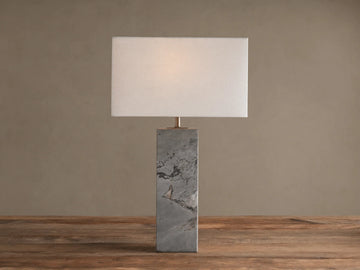 Oren Table Lamp: Rectangular white linen shade over a gray stone base with dark veining, sitting on a rustic wooden table.