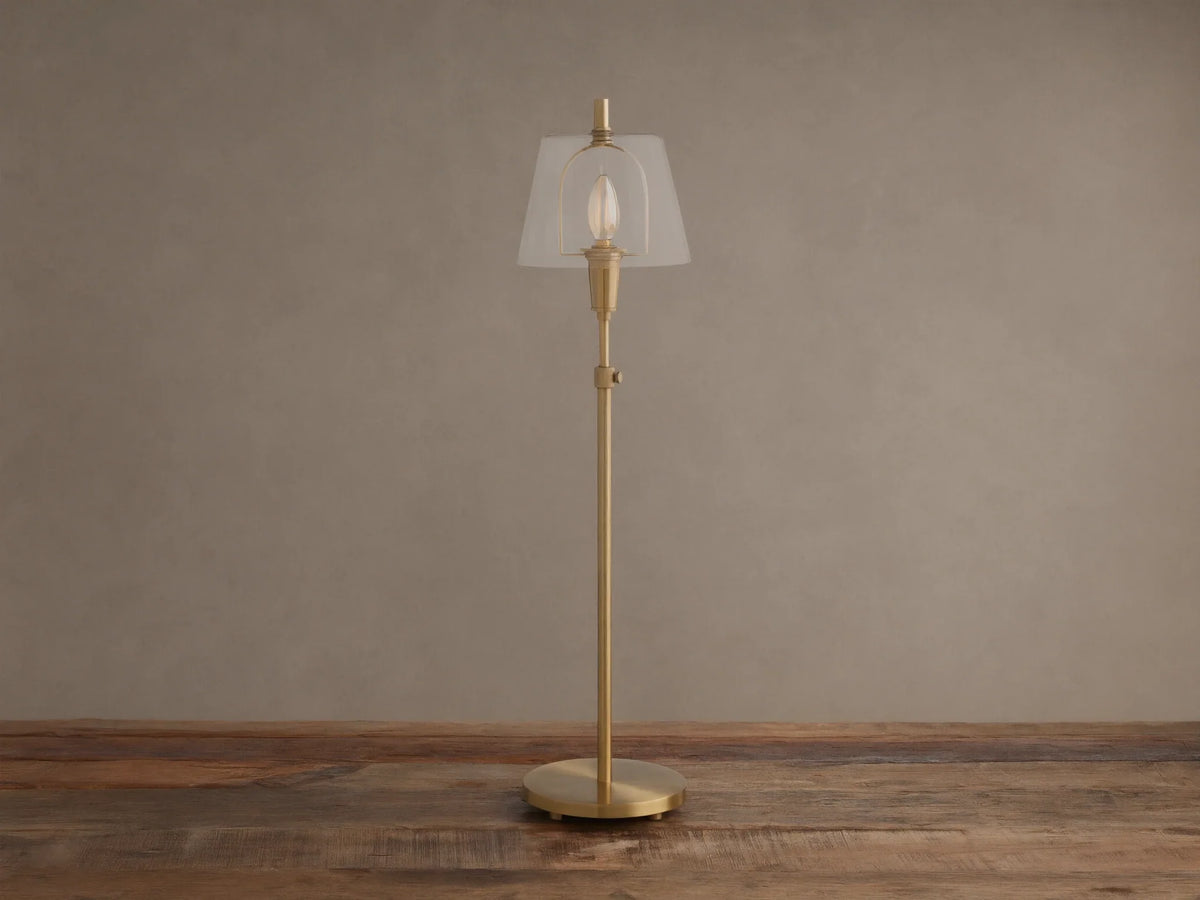 Brass Candice Table Lamp with a clear conical shade and illuminated bulb, on a wooden surface against a textured beige wall.