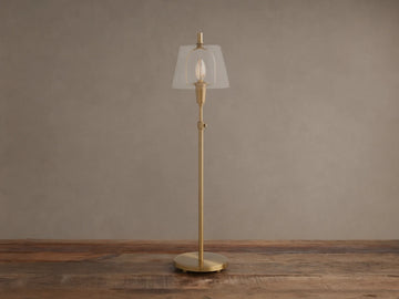 Brass Candice Table Lamp with a clear conical shade and illuminated bulb, on a wooden surface against a textured beige wall.
