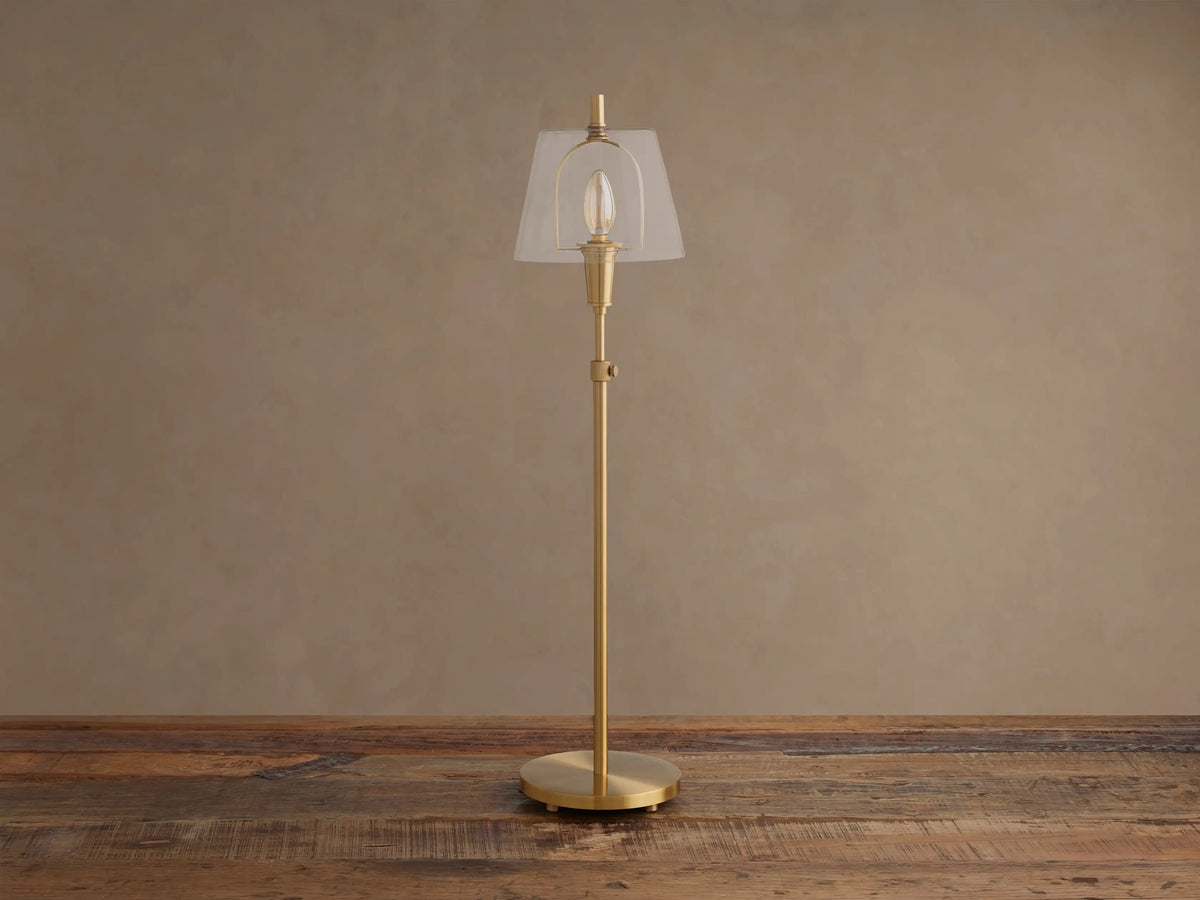 Brass Candice Table Lamp with a clear conical shade and illuminated bulb, on a wooden surface against a textured beige wall.