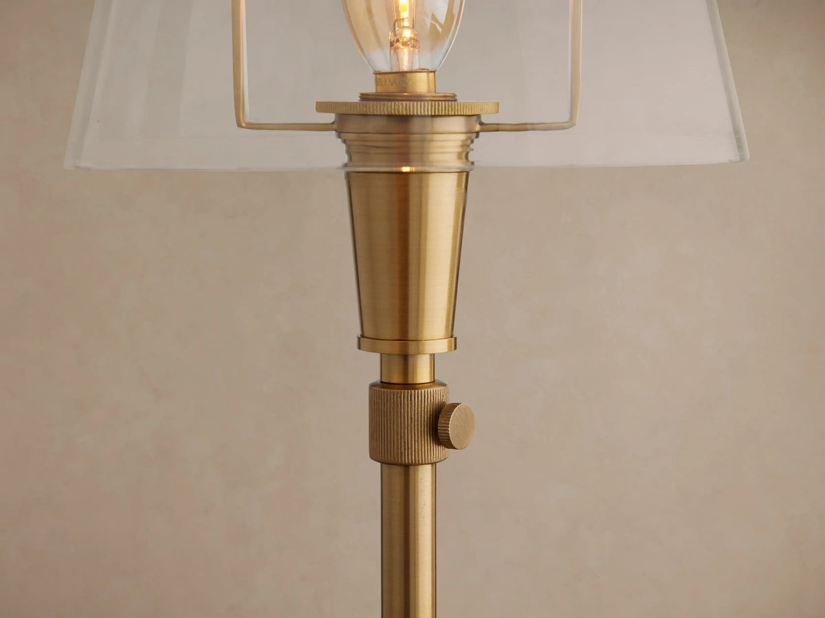 Candice Table Lamp in brass finish with a clear shade and exposed bulb, standing on a wooden floor against a beige wall.