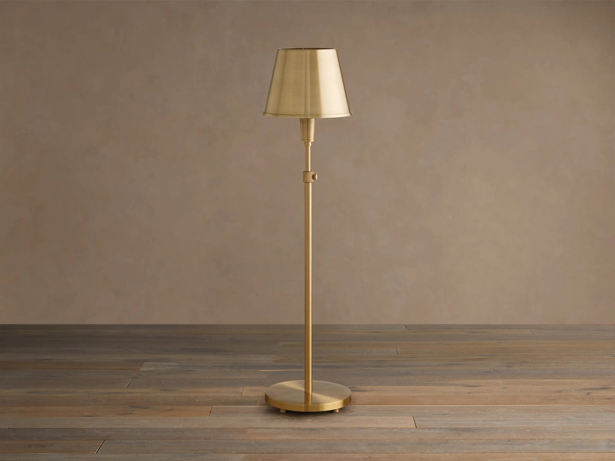Illuminated Candice Table Lamp in brass, with a cone-shaped brass shade, standing on a wooden floor against a beige wall.