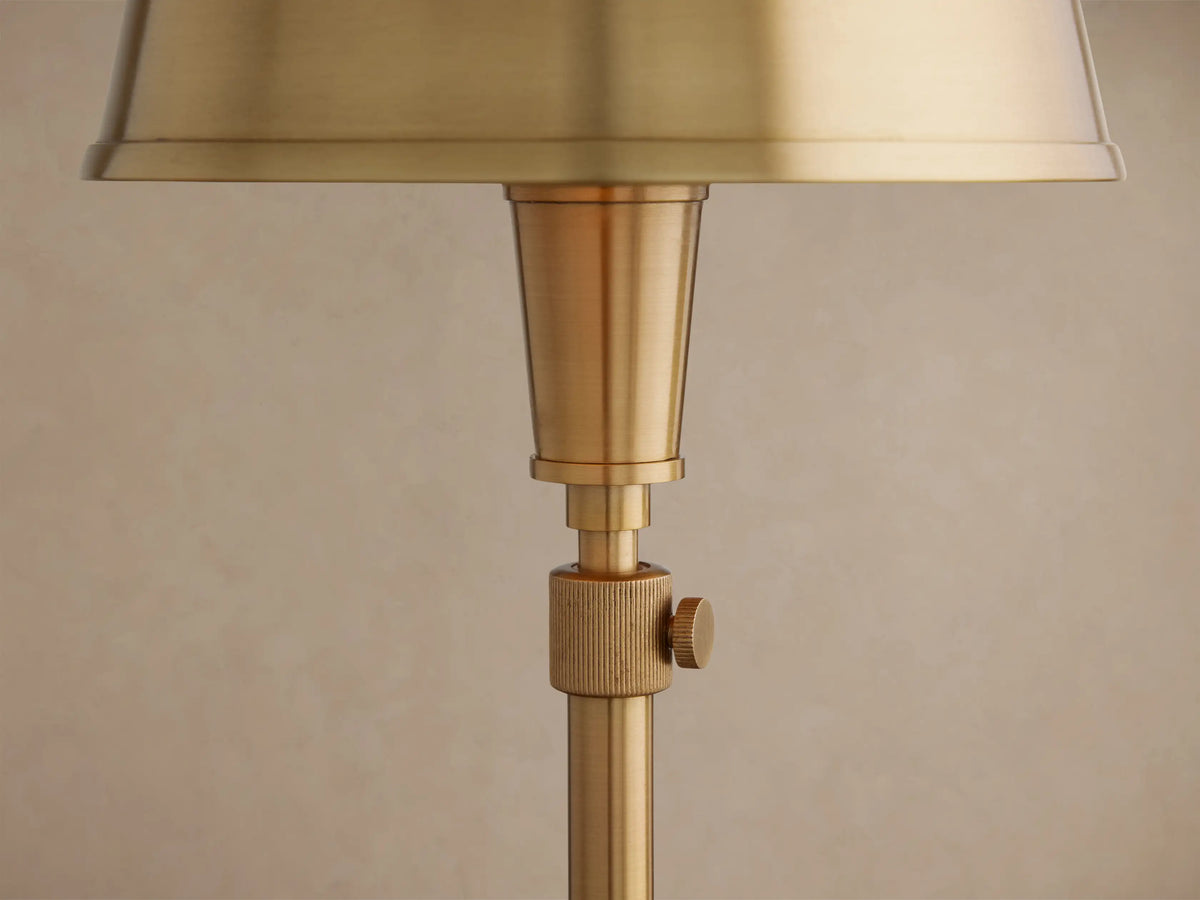 Candice Table Lamp. Tall, slender brass lamp with a tapered brass shade, slim stem, and round base, standing on a wooden floor.
