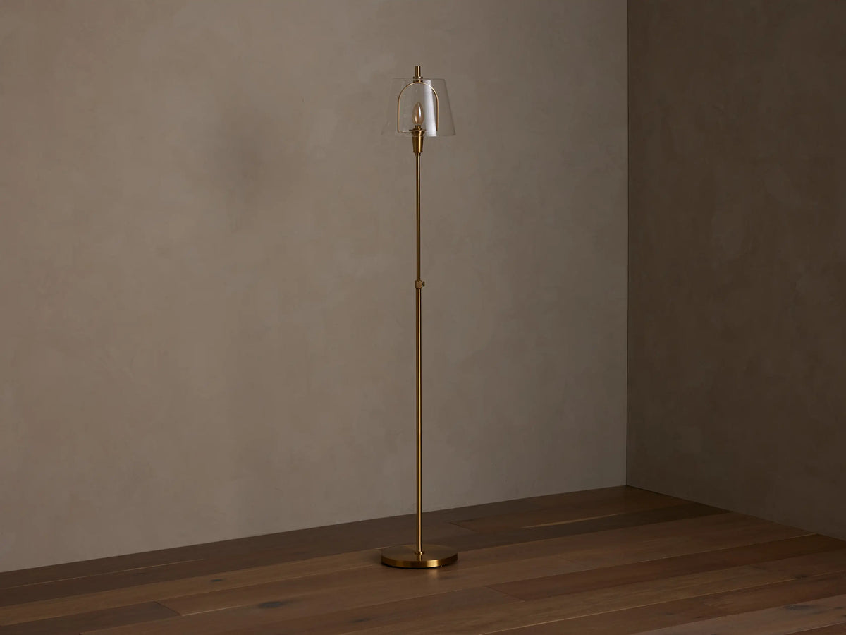 Candice Floor Lamp with a brass finish and a clear bell-shaped glass shade, emitting warm light in a dimly lit room.