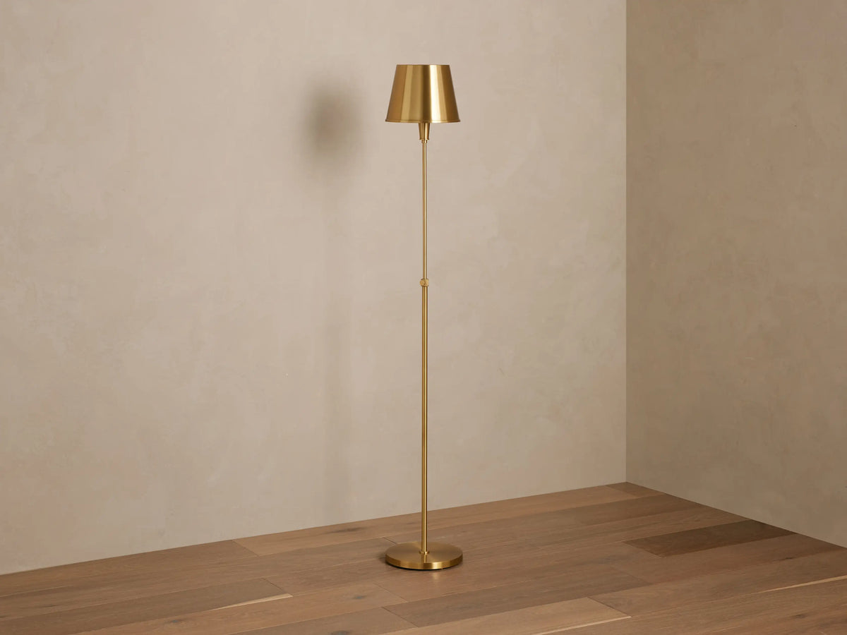 Candice Floor Lamp in brass finish with a tapered shade, lit and casting a warm glow, standing in a room corner on a wooden floor.