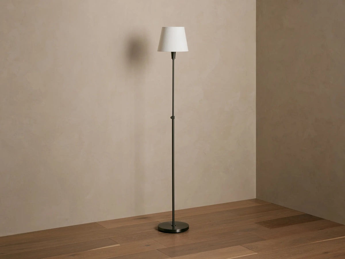 Candice Floor Lamp with dark pewter stand, white tapered shade emitting warm light, against a beige wall and wood floor.