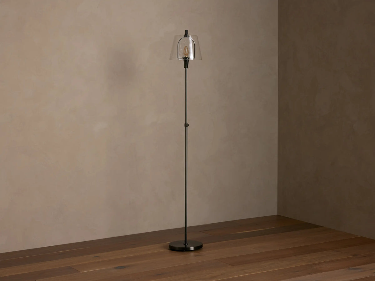 Candice Floor Lamp in dark pewter with a clear glass shade and glowing bulb, standing in a room corner with brown walls and wood floor.