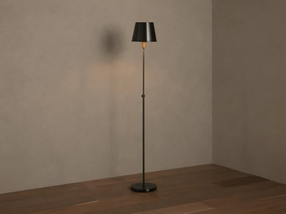 Candice Floor Lamp with a slender dark pewter-finished stand & tapered shade, illuminated, in a room corner with wood floor.