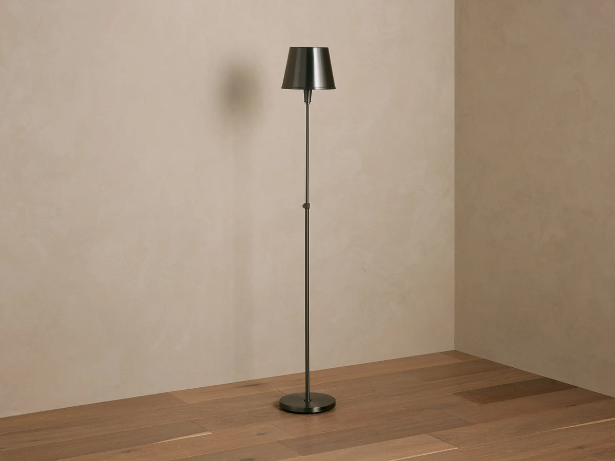 Candice Floor Lamp with a slender dark pewter-finished stand & tapered shade, illuminated, in a room corner with wood floor.
