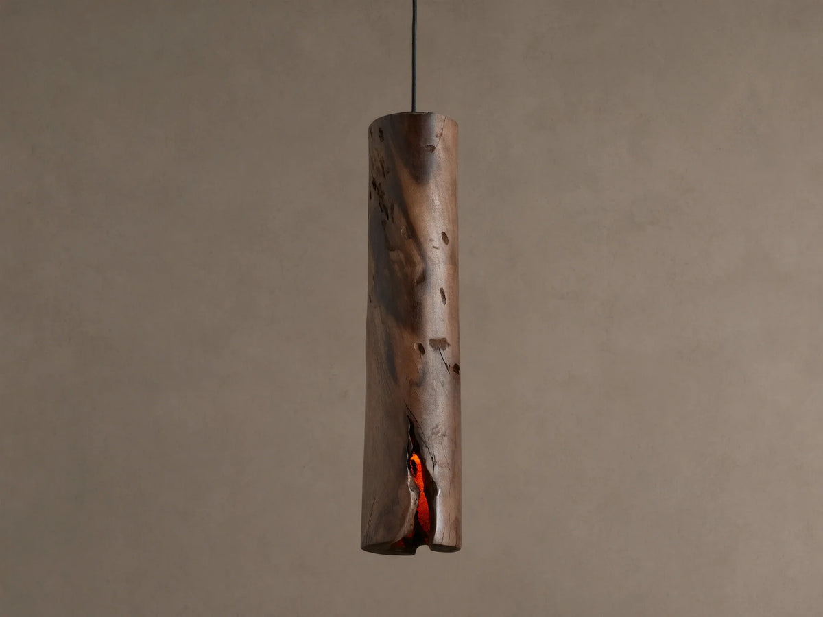 Dedalo Pendant, a cylindrical milpa wood light with natural texture, hangs vertically. An orange glow emanates from a front crack.