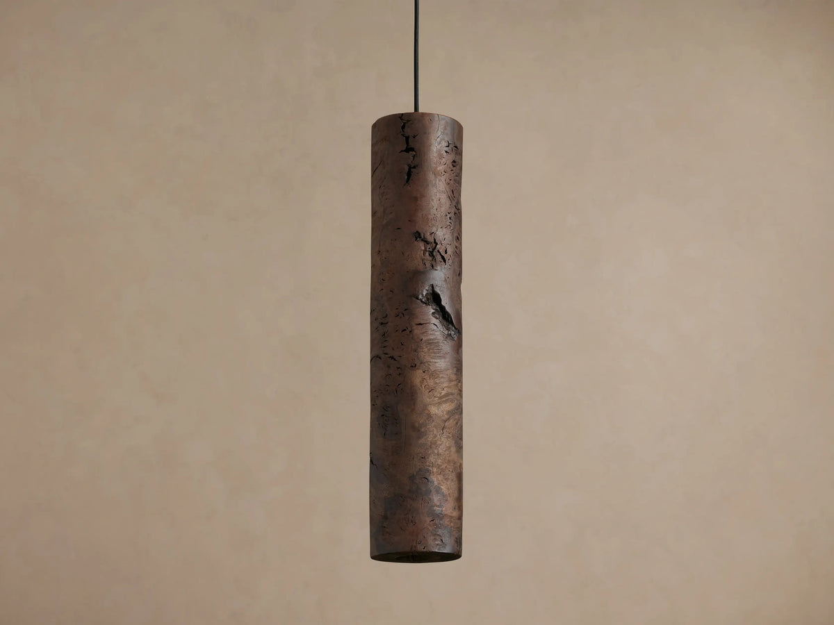 Three cylindrical Dedalo Pendants of dark milpa wood hang, natural cracks emitting warm orange light against a light wall.