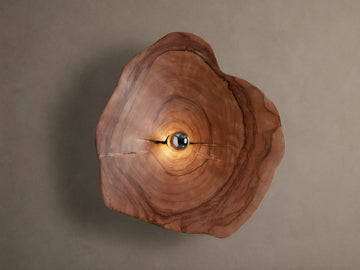 Arruga Sconce: Irregular natural wood slice with visible grain and cracks, light bulb glowing warmly in the center, mounted on a neutral wall.